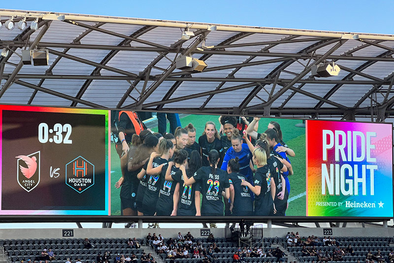 ACFC's team with Press in the front is on the big screen in their Pride jerseys. On one side of the team, the clock counts down to kick off. On the other, the screen says "Pride Night."