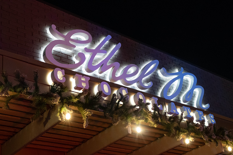 The Ethel M Chocolates sign over the factory shop. The sign is hued in purple and blue, and the overhang below has white lights and is decorated with holly.
