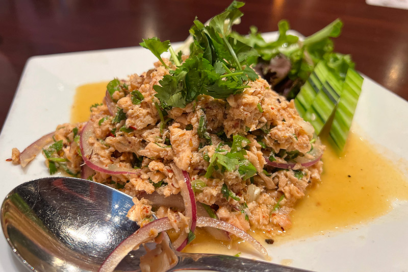 A close-up of a dish of salmon larb. A pile of roughly chopped salmon is mixed with spices and garnished with parsley, red onions and cucumbers.