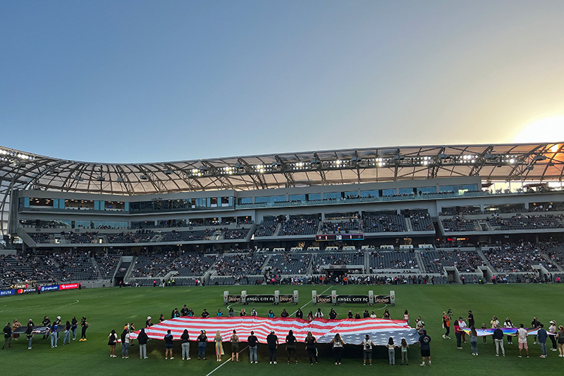 A large American flag and pride flags are held open on the field at the Banc of California. The far side of the stadium meets the sunset sky in the background.