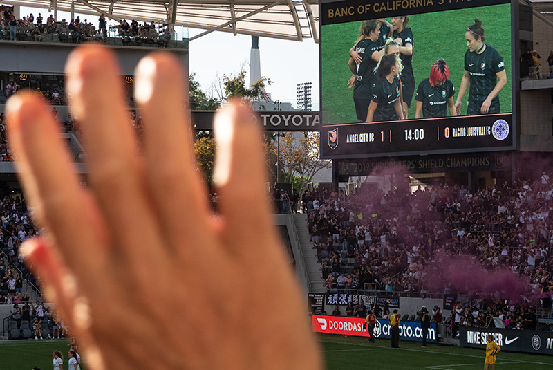 A fan's hand dominates the foreground. In the background on a screen, ACFC players celebrate a goal.