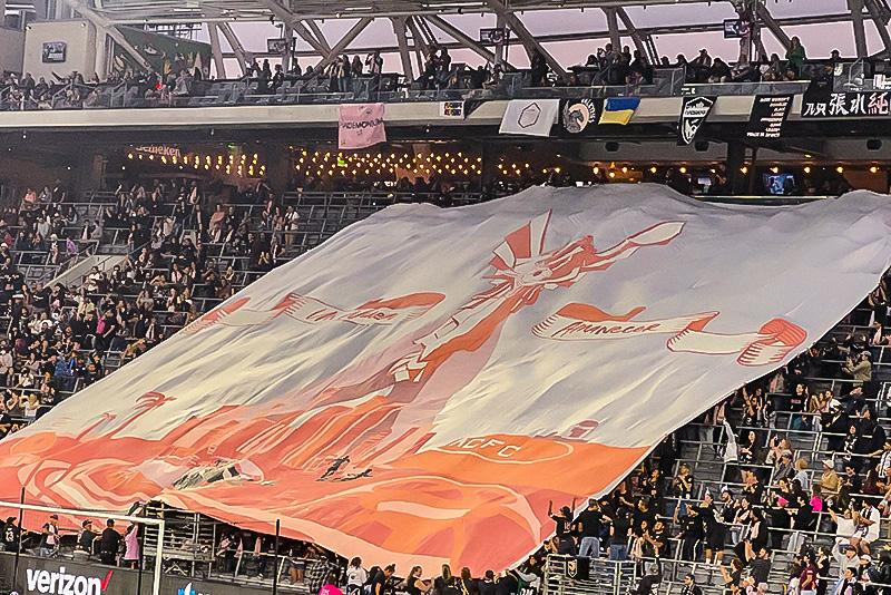 The inaugural Angel City tifo unfurls over fans in the supporter section at Banc of California. The tifo is a pink angel a pink cityscape of LA on a white background.