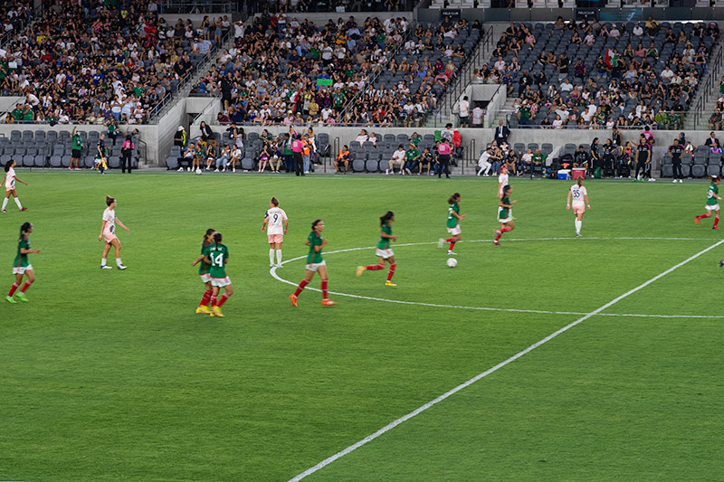 The Mexican National team runs forward on the pitch. Angel City players dot the field between them. In the background, fans look on from the stands.