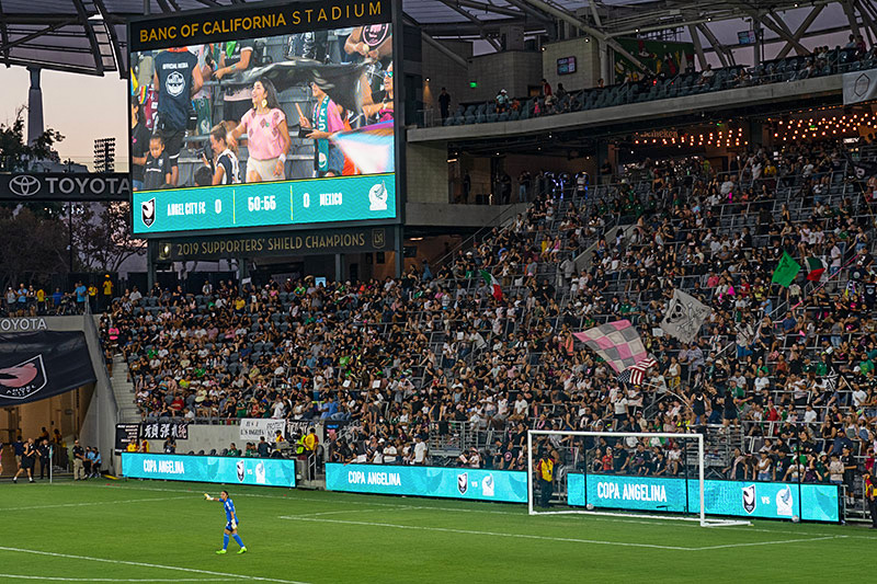 Fans cheer in the supporter section. In front of them, a screen shows a fan close up and screens display "Copa Angelina"