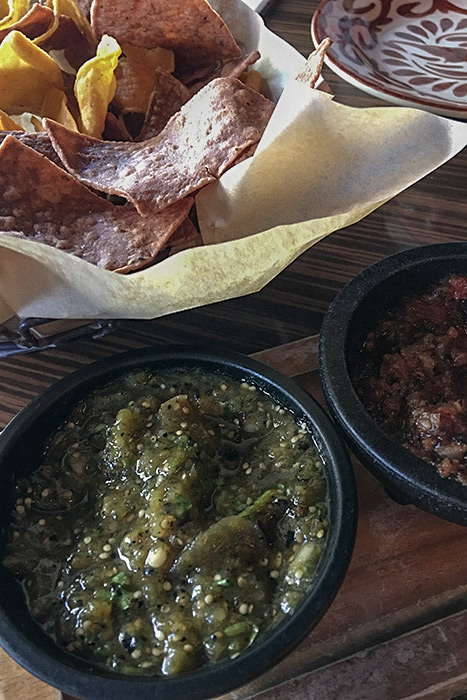 A bowl of salsa verde sits in the forefront on a wooden tray. To its right is a bowl of red fire-roasted salsa. In the background, a basket of purple and yellow tortilla chips.