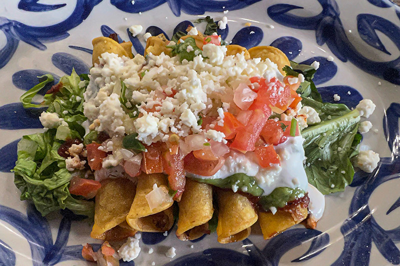 Five beef taquitos are topped with guacamole, queso fresco and pico de gallo on a blue and white plate.