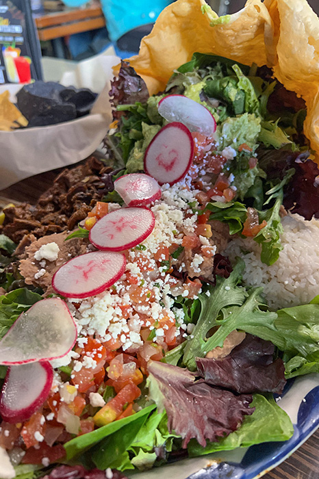 A close-up of a huge taco salad. The shell bowl is tilted on its side with the salad spilling out of it in a neat line. You can see a bed of lettuce with a pile of white rice and pile of refried beans on one side and a pile of beef on the other side. The lettuce is topped with pico de gallo, queso fresco, and radishes.