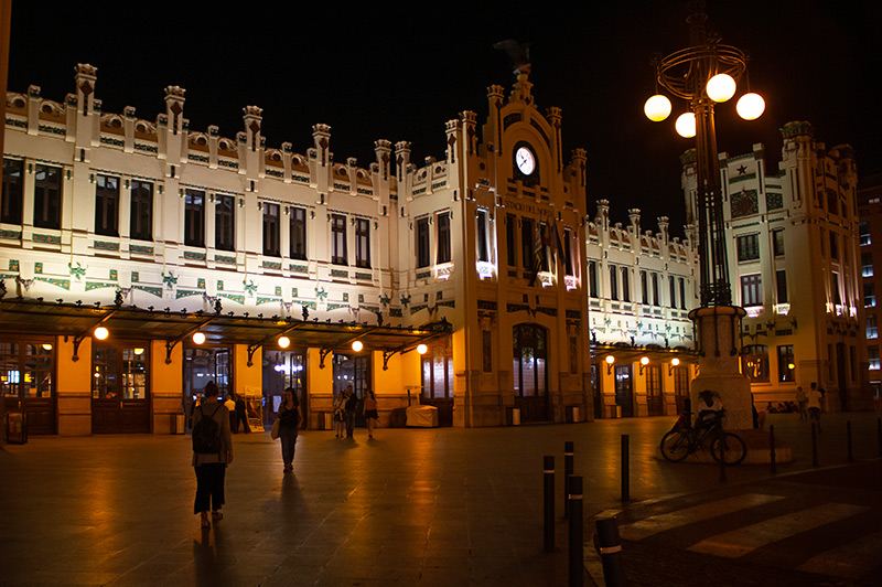 Estacio del Nord in Valencia Spain at night. The building looks two-toned yellow in the darkness. A very pale yellow or cream avoe a glass awning that hangs over the doors and deeper yellow on the walls next tot eh doors. Light fixtures with large round bulbs hang on the walls between the doors of hte station. In the center, there is a protruding section of the station that sits further forward than the rest of the station. It has an arched top with small decorative turrets rising around it and a clock in the face above an ornate door. The decorative turrets stretch along the entire roof at regular intervals. The upper walls of the building has regular sets of windows. The wall below has decorative mosaics in a pattern.