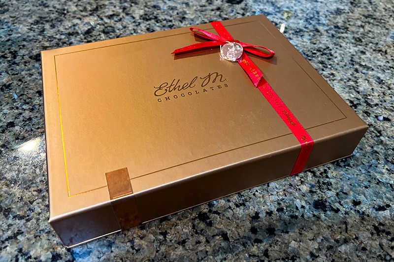 The chocolate box from Ethel M sits on a marble counter. The box is gold with Ethel M printed on it and a red holiday ribbon.