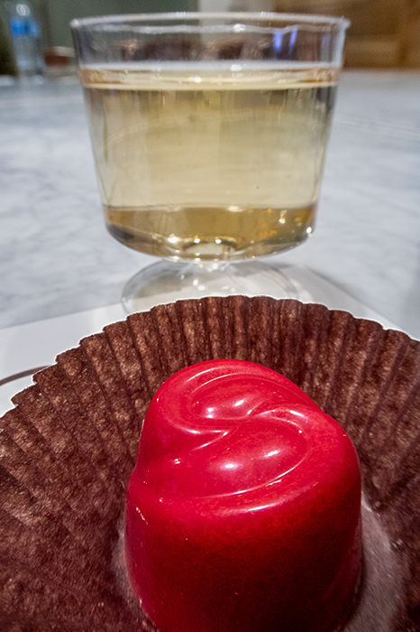 A close-up of the peppermint silk truffle at the Ethel M tasting. The truffle is colored bright red and sits in a brown paper. The cup of champagne dominates the background.