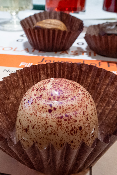 A close-up of the white chocolate vanilla truffle at the Ethel M tasting. The truffle is white with purple-red flecks. Other truffles and small cups of wine are visible in the background.