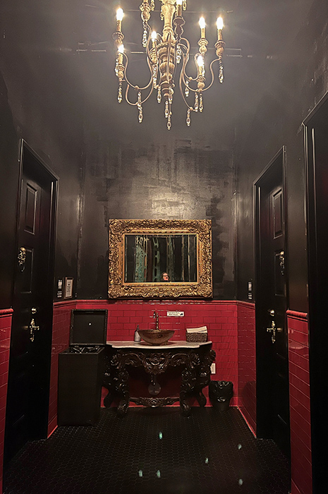 An ornate golden mirror hangs over a sink basin, a gold chandelier hangs above, doors are on either side to individual toilet stalls in the bathroom area of Lost Spirits distillery.