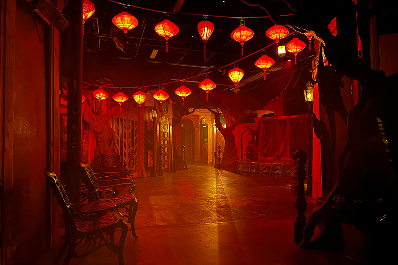 An "alley" inside Lost Spirits distillery. To the left, metals chairs line the wall. Chinese lanterns cross the "alleyway". Leafless tree cutouts with detailed trunks appear along the alley, along with intricate doors. The entire scene is softly tinged in a deep red glow.