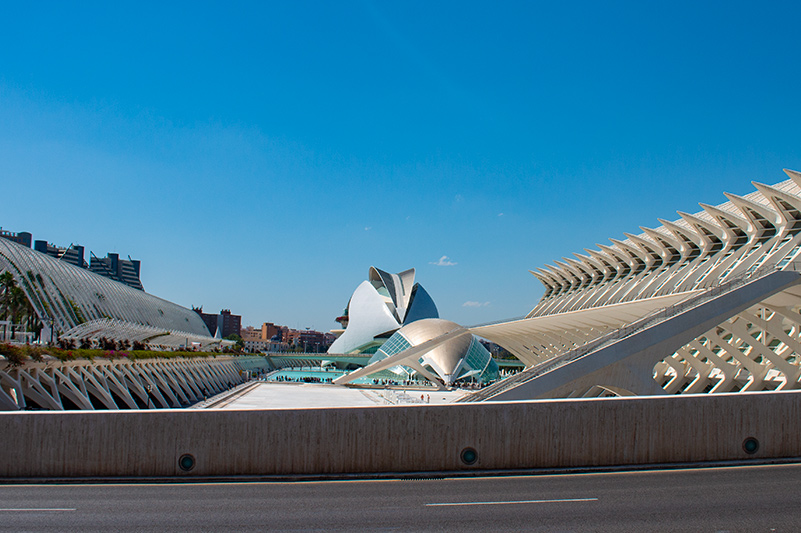 Parc del Museu de les Ciències in Valencia, Spain. To the left, a long white structure with arches at the bottom and alternating arches and open spaces at the top sits stretches back beside a concrete plaza with two buildings. To the right, a long structure with v-shaped buttresses that create a sort of makeshift awning and jutting windows that look somewhat like spikes stretches along the other side of the plaza. The two buildings on the plaza sit somewhat back with the concrete plaza in front. The front building is shaped like a half egg and has green-tinted windows on both sides and a gold panel down the middle. The back building is taller and shaped somewhat like a helmet. The sides are white and slightly separate from the center, which is metallic and sits slightly above the rest of the building.