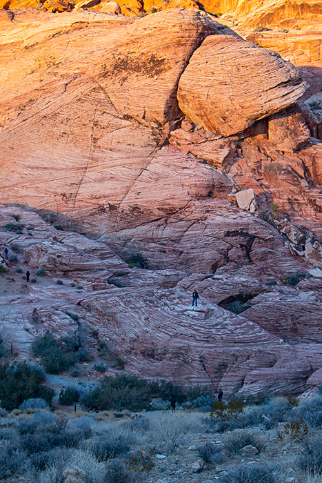 Sandy brushland sits in shadows in the foreground. Behind it, the shadows rise halfway up red striped boulders, while the top of the boulders shine in the sun.