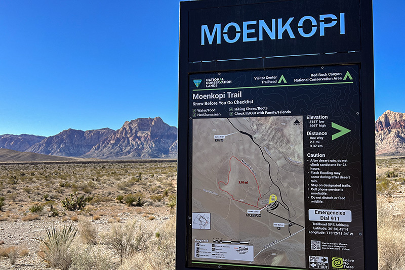 The trailhead sign for Moenkopi Trail. The trail name is in cut-out letters at the top of the sign with the blue sky showing through. Below that, the trail map and information is on display. In the background, the mountains loom in the distance.