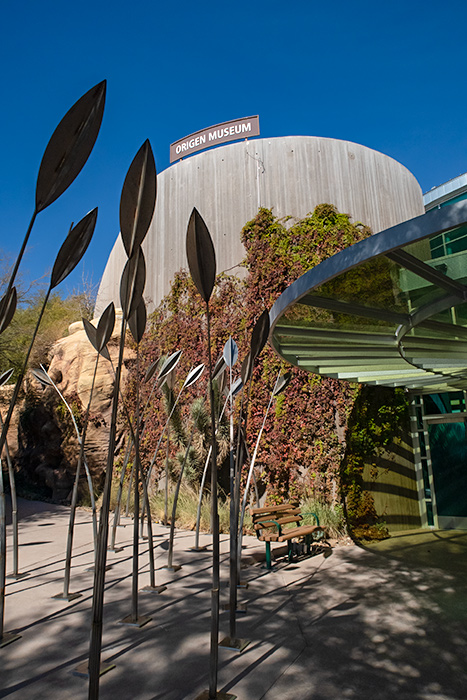 The outside of the Origen Museum at Springs Preserve. A tall concrete silo-like column rises into the sky with a brown Origen Museum sign sitting atop it. Ivy climbs up the wall and sculptures of leaves stand in front of it. To the right, a see-through, green-tinted awning stands over the entrance doors, and to the left there's a cave-like passageway.