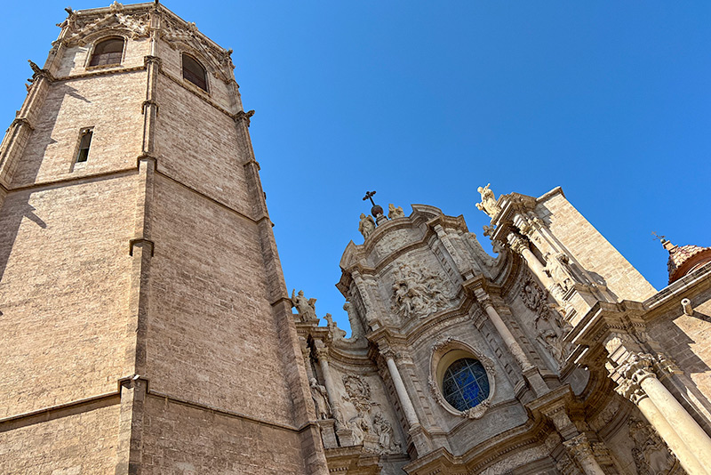 The upper portion of Valencia cathedral. To the left is a brick tower with an ornate design and windows at the top that rises higher than the decorative wall above the cathedral entrance. The wall is curved inward and has many ornamentations. At the top there is a cross and statues and columns are visible, though none are large or close enough to be made out in detail.