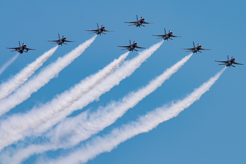 Thunderbirds fly in formation over Las Vegas. Eight planes fly in a V, one in the front, one trailing directly behind it and the other six fanning out in wings on either side. The sky is blue, and white contrails fill the screen behind the jets.