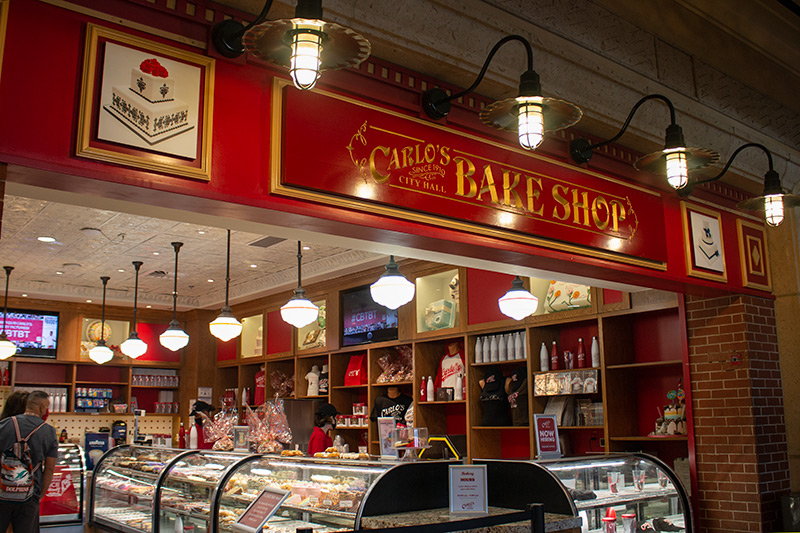 the exterior of Carlo's Bake Shop, the sign for the bake shop at top, display cases of baked goods in front, merch on shelves in the back