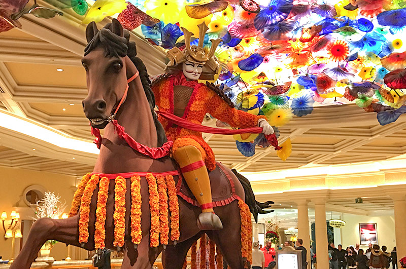 a display of an asian soldier riding a horse sits under Chihuly's Fiori di Como display - a display of multi-colored glass in yellows, reds and blues mounted in the ceiling in the lobby of the Bellagio hotel