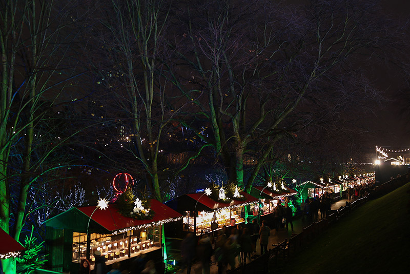 Red-roofed Christmas market stalls line the walkway through Princes Street Gardens. White-lit star decorations rise between them. People fill the walkway. Behind the stalls, the naked-branched trees are lighted in green as they stand against a dark sky.