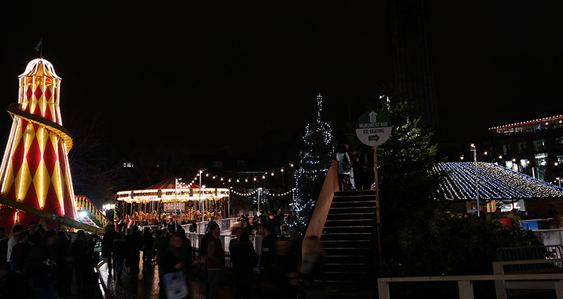 The smaller holiday set-up in St. Andrew Square. To the left, a tall old-school slide with a red and gold diamond pattern stands. Further back to the right, a carousel is lighted, and to the right stands the ice skating rink and look-over bridge built above it.