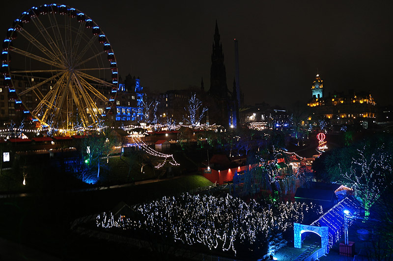 Princes Street Gardens in Edinburgh decorated for the holidays. To the right, a small, temporary train tunnel is lighted bright blue with a small track running through it. Behind that, shrubbery decked out in white lights leads to the red houses of Santa's Village. Slightly elevated above that on the left is a line of red-topped vendor stalls and the large ferris wheel lighted in blue sits behind it. In the far distance, a striking building is lighted in light green and gold.