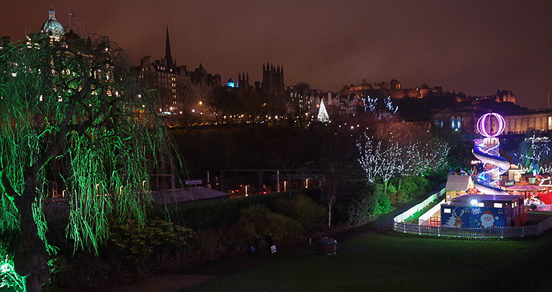 The distinctive outline of Old Town Edinburgh looms in the background over Santa Land in Edinburgh's Princes Street Gardens. From left to right, the green dome of the Bank of Scotland, a spire, and the battlements of a medieval building lead on to Edinburgh Castle. At the front left, a green-lit weeping willow dominates, while on the right, a brightly-lit slide with a ball on top dominates Santa's playland.